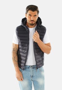 Gilet trapuntato blu con cappuccio, chiusura con zip e due tasche laterali. Indossato sopra una t-shirt bianca e jeans azzurri. Texture liscia e slanciata.