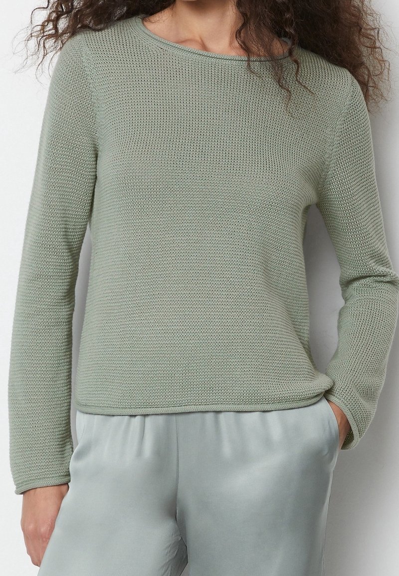 Femme portant un pull en maille vert sauge à manches longues et un pantalon en satin vert clair, debout avec une main dans la poche devant un fond uni.