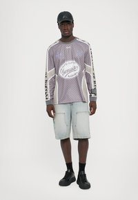 Langarmige grijze shirt met patroon en graphics, gecombineerd met lichtblauwe denimshorts. Zwarte sneakers en een pet maken de outfit compleet.