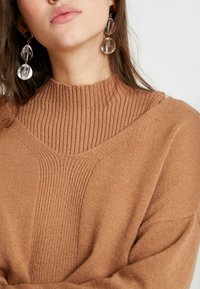 Maglione lavorato a maglia marrone con collo alto a coste e una vestibilità comoda. Orecchini pendenti trasparenti aggiungono un elemento di contrasto.