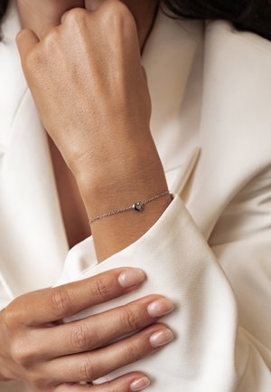 Close-up of a wrist wearing a delicate silver bracelet with a small heart charm, against a cream-colored blazer sleeve.