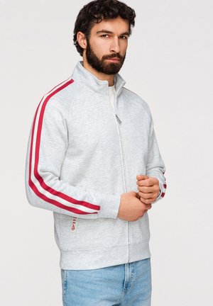 Hombre con cabello oscuro y rizado y barba, que lleva una chaqueta gris clara con cremallera y rayas rojas en las mangas y vaqueros azules, de pie frente a un fondo liso.