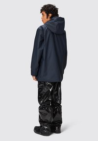 Giacca impermeabile blu navy con cappuccio, caratterizzata da una vestibilità rilassata. Abbinata a pantaloni neri lucidi con fondo arricciato. Sfondo semplice.