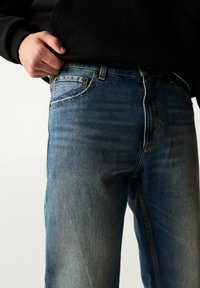 Jeans de mezclilla azul oscuro con un corte clásico, con cierre de cremallera, diseño de cinco bolsillos y un sutil desgaste en los muslos. Usados con una sudadera negra con capucha.