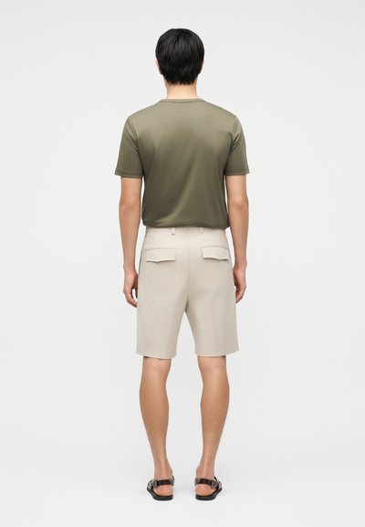 Άνδρας που στέκεται γυρισμένος προς τα πίσω φορώντας μια ελιάς πράσινη μπλούζα, μπεζ σορτς μέχρι το γόνατο με πίσω τσέπες με καπάκι και μαύρα σανδάλια.