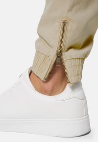 Pantalons beige avec une fermeture éclair à la cheville, dotés de poignets élastiques. Portés avec une paire de baskets blanches. Texture de tissu lisse.