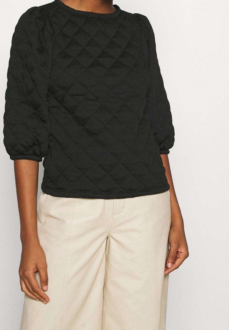 Svart vadderad pullover med rund halsringning och puffärmar. Tyget har ett diamantsmönster och är kombinerat med ljusbeige vida byxor.