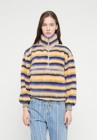 Lun, busket pulloverjakke med horisontale striber i gul, lilla, grå og sort. Lynlås ved kraven og ballonsærmer. Båret med blå jeans med striber.