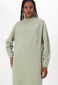 Robe tricotée vert clair avec un col montant, des manches longues et des poignets côtelés, présentant une texture douce et une coupe décontractée.