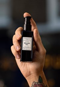 Bottiglia nera di vetro con dispenser a pompa; etichettata "MURDOCK REDCHURCH BEARD OIL"; capacità 50 ml. Mano che tiene la bottiglia con un tatuaggio visibile.