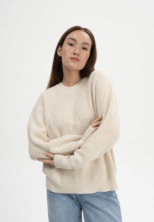Cremfarbener Strickpullover mit strukturiertem Muster, rundem Rundhalsausschnitt und überschnittenen Schultern, kombiniert mit hellblauen Jeans.