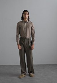 Bărbat cu păr lung până la umeri, de culoare închisă, îmbrăcat cu o cămașă cu nasturi de culoare taupe, pantaloni pliți de culoare măslinie închisă și mocasini bej, stând pe un podea gri.