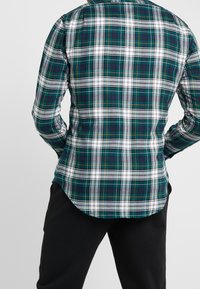 Zielono-biała koszula w kratę z ciemnoniebieskimi akcentami, długimi rękawami i zaokrąglonym dołem. Materiał wygląda na gładki, z klasycznym wzorem w kratę.