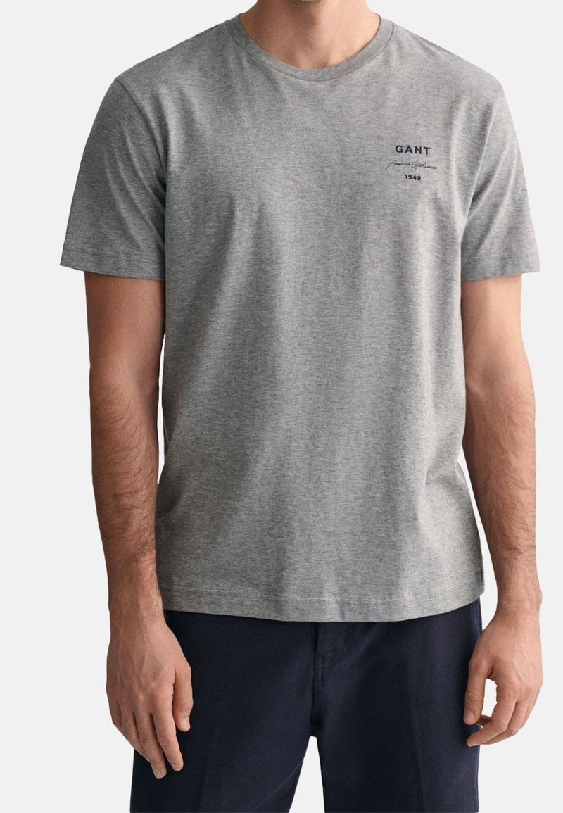 Homme portant un T-shirt gris clair à manches courtes avec le texte « GANT American Sportswear 1949 » sur la poitrine et un pantalon foncé.