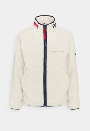 Witte fleecejas met marineblauwe rits, rood trekkoordje, hoge kraag met gedeeltelijke rode en zwarte tekst, borstzak en zijzakken.