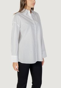 Camicia bianca con bottoni, maniche lunghe, caratterizzata da un sottile motivo testurizzato, colletto e orlo curvo. Abbinata a pantaloni neri.