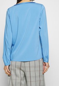 Donna che indossa una camicia a maniche lunghe blu chiaro e pantaloni grigi a quadri, vista da dietro su uno sfondo bianco semplice.