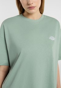 T-shirt en coton vert clair avec un col rond, des manches courtes, et un petit logo blanc sur la poitrine. Texture lisse, coupe décontractée.