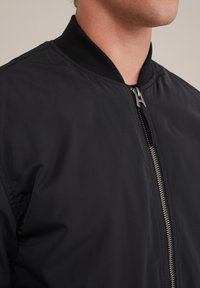 Černá bomber bunda s žebrovaným límcem, předním zipem a hladkou texturou látky. Disponuje minimalistickým designem a čistými liniemi.