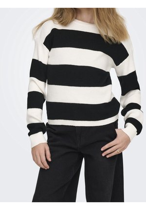 Kvinde iført en langærmet sort-hvid sweater med horisontale striber og sorte bukser, med den ene hånd i lommen og en ring på den anden hånd.