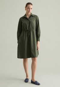 Kleider in khakigrün mit Knopfverschluss, tailliertem Gürtel, langen Ärmeln und einem dezenten Texturmuster. Kombiniert mit navyblauen, gemusterten Slippern.