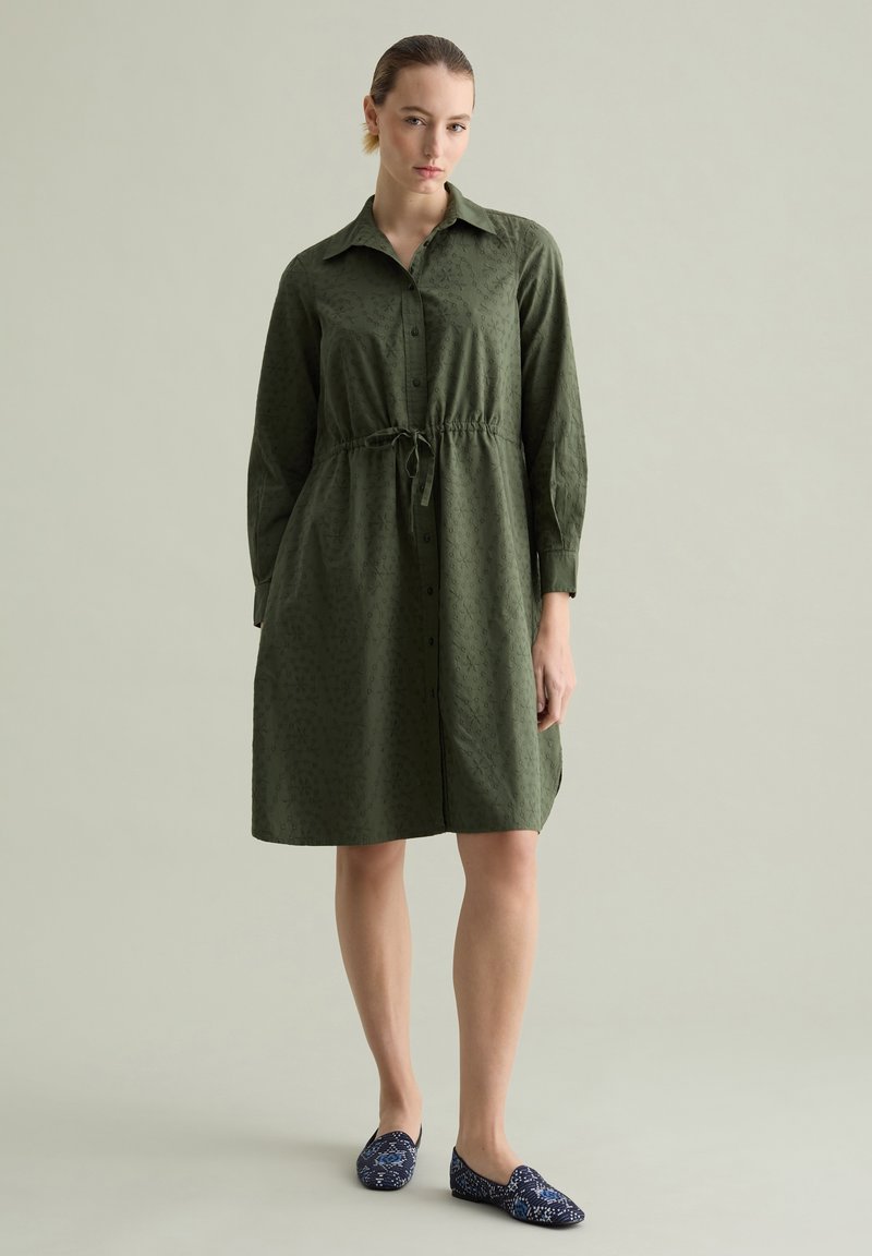 Kleider in khakigrün mit Knopfverschluss, tailliertem Gürtel, langen Ärmeln und einem dezenten Texturmuster. Kombiniert mit navyblauen, gemusterten Slippern.