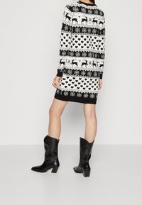 Robe-pull à motifs noirs et blancs avec des rennes, des cœurs et des flocons de neige ; manches longues et poignets côtelés. Portée avec des bottes noires à talons.