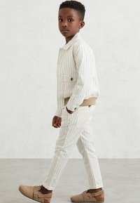 Bela stripe jakna in hlačne kompleta z bež akcenti. Material se zdi lahek in teksturiran. Obutev je bež barve s pasom in ravnim podplatom.