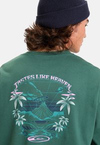 Groene sweatshirt met grafisch ontwerp dat tropische motieven, rook en een vulkaan bevat. De tekst luidt "Smaakt Naar de Hemel" in het roze.