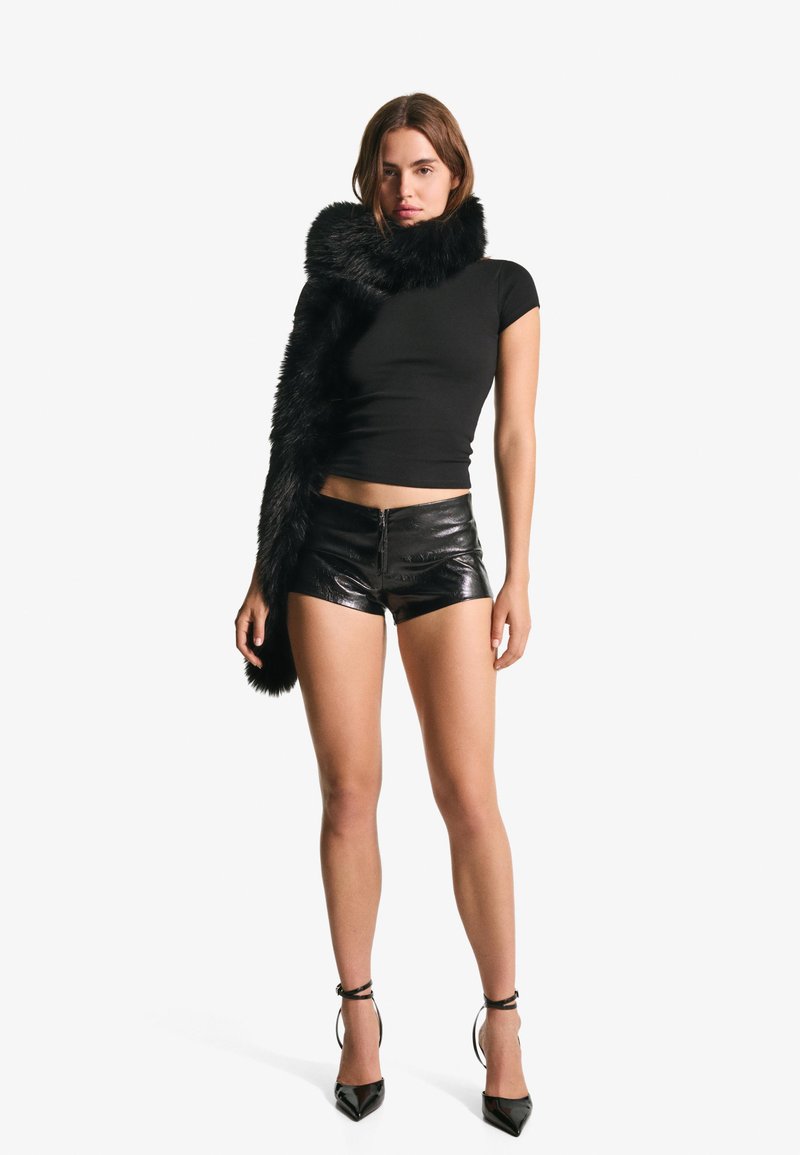 Čierny priliehavý top s krátkym rukávom skombinovaný s lesklými čiernymi koženými šortkami a dlhým, nadýchaným čiernym šálom; model má na sebe čierne lodičky s špicatou špičkou.