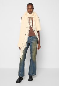 Crème gebreide sjaal met franjes, gedragen over een bruin geribbeld topje en gecombineerd met blauwe flared jeans en zwarte platformlaarzen.