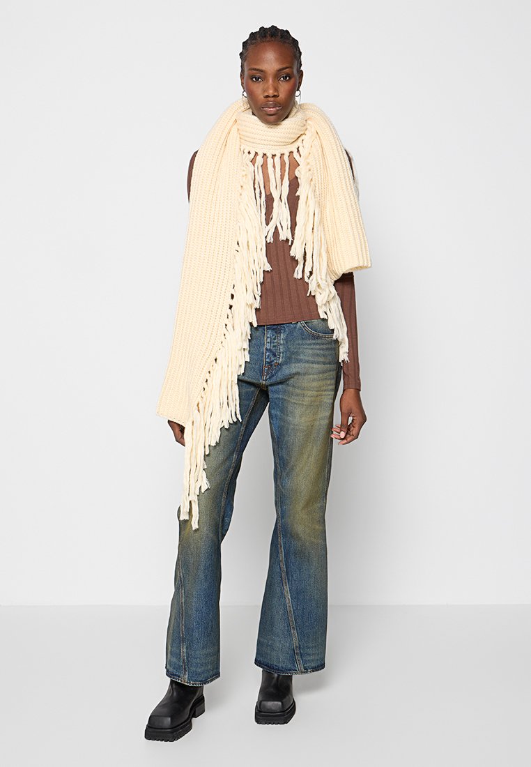 Crème gebreide sjaal met franjes, gedragen over een bruin geribbeld topje en gecombineerd met blauwe flared jeans en zwarte platformlaarzen.