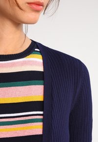 Laivastonsininen ribbineulottu neuletakki, joka peittää monivärisen raidallisen neuleen, jossa on vaaleanpunaisia, vihreitä, keltaisia ja laivastonsinisiä raitoja ja teksturoitu neulos.