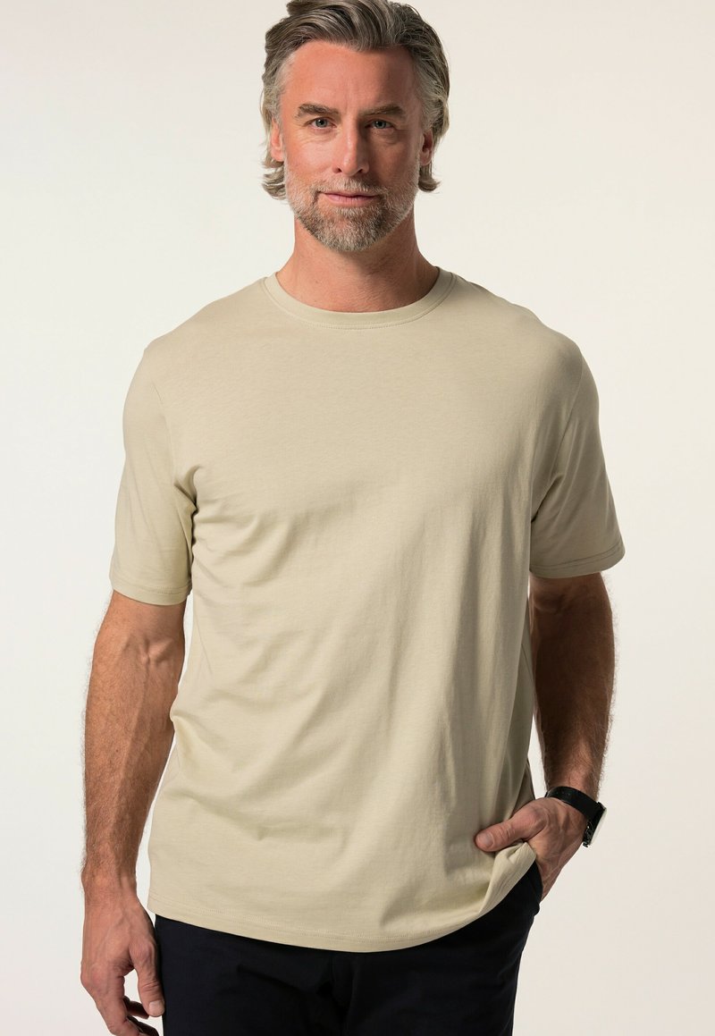 Beige T-Shirt mit kurzen Ärmeln aus glattem Stoff, mit einem runden Halsausschnitt und einer lockeren Passform. Getragen mit dunklen Hosen und einer Uhr.
