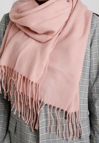 Écharpe rose pâle à franges posée sur un blazer gris à carreaux, couvrant la partie supérieure de la poitrine et du cou.