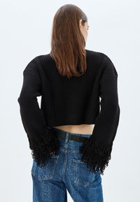 Schwarzer, taillierter Pullover mit gerippter Textur, weiten Ärmeln, die mit langen Fransen verziert sind, kombiniert mit hoch taillierten blauen Jeans.
