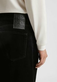 I pantaloni di jeans neri presentano una toppa in pelle nera texture sul retro, con cuciture bianche e un design slim fit. Indossano una maglietta bianca a maniche lunghe.