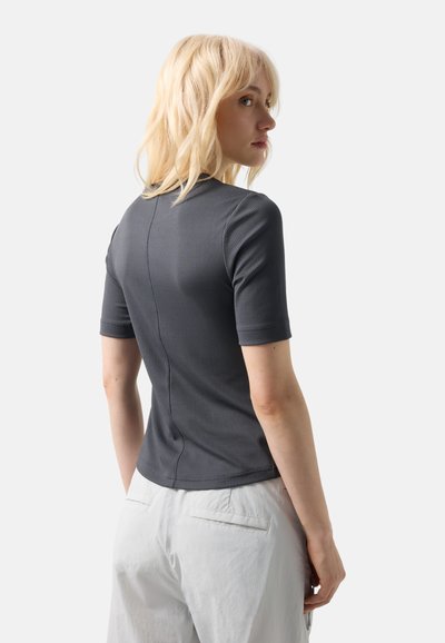 Kvinde med blondt hår iført en tætsiddende, mørkegrå, ribstrikket top med korte ærmer og hvide bukser, der kigger over sin skulder mod højre.