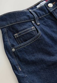 Jean en denim bleu foncé avec des coutures contrastantes claires, une poche avant et un bouton en métal. La texture semble lisse et solide.