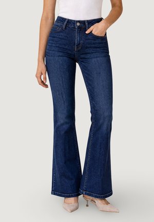 Persona che indossa jeans a vita alta a zampa di colore blu scuro e décolleté beige con punta a punta con una canottiera bianca.