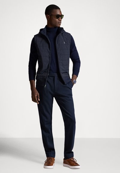 Vyras dėvi tamsius akinius nuo saulės, tamsiai mėlyną pūstą liemenę ant tamsiai mėlyno golfo, tamsiai mėlynas kelnes, rudas sportbačius ir laikrodį riešo, stovintis su viena ranka kišenėje.