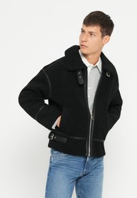 Schwarze Fleecejacke mit breitem Kragen, Reißverschluss und Lederakzenten. Sie verfügt über zwei Seitentaschen und eine strukturierte Oberfläche.
