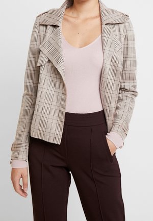 Woman wearing a beige plaid jacket over a light pink ribbed tank top and dark brown high-waisted pants with one hand in pocket.