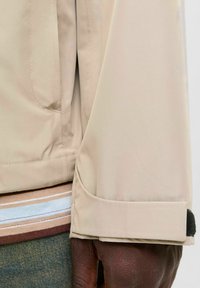 Beige vanntett jakkesleeve med en sikret mansjett, med en svart borrelåsdetalj og en synlig lomme-søm. Stripete fôrdel av foret vises delvis.