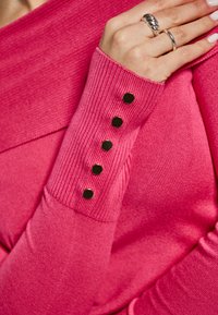 Top rosa a maniche lunghe con costine, dal collo ampio e dettagli con bottoni sulle maniche. Texture morbida e design aderente.