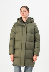 Vrouw in een olijfgroene gewatteerde pufferjas met capuchon, staande tegen een effen witte achtergrond, met handen ontspannen langs haar zij.