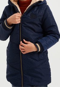 Dunkelblauer Puffer-Jacke mit Leopardenmuster, ausgestattet mit einer gefütterten Kapuze aus Kunstfell und gerippten Bündchen. Mit Reißverschluss vorne und Seitentaschen.