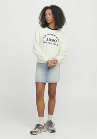 Felpa chiara con testo blu navy, abbinata a una mini gonna di jeans. Presenta sneakers grigie e capelli ricci e texturizzati.