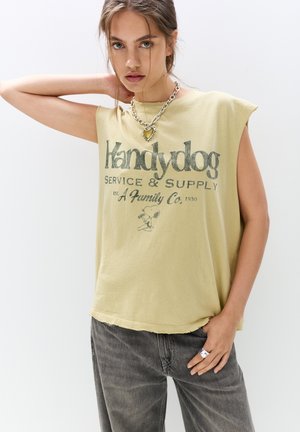 Olivgrünes ärmelloses T-Shirt mit einem ausgefransten Saum, versehen mit schwarzem Text "Handydog Service & Supply" und einer Cartoon-Grafik. Getragen mit grauen Jeans.