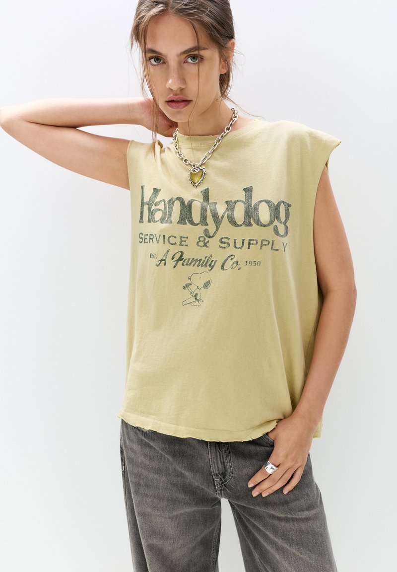 Oliven grøn, uden ærmer t-shirt med en slidt kant, prydet med sort tekst "Handydog Service & Supply" og en tegneserie-grafik. Båren med grå jeans.